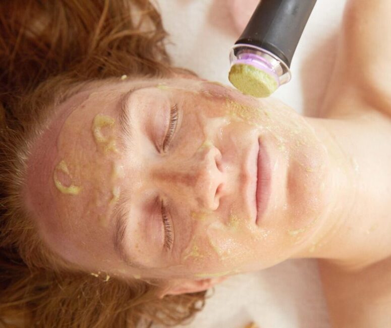 Woman with red hair lays down with eyes closed and gold facial product on her skin. Wand from facial machine shows in top left corner.
